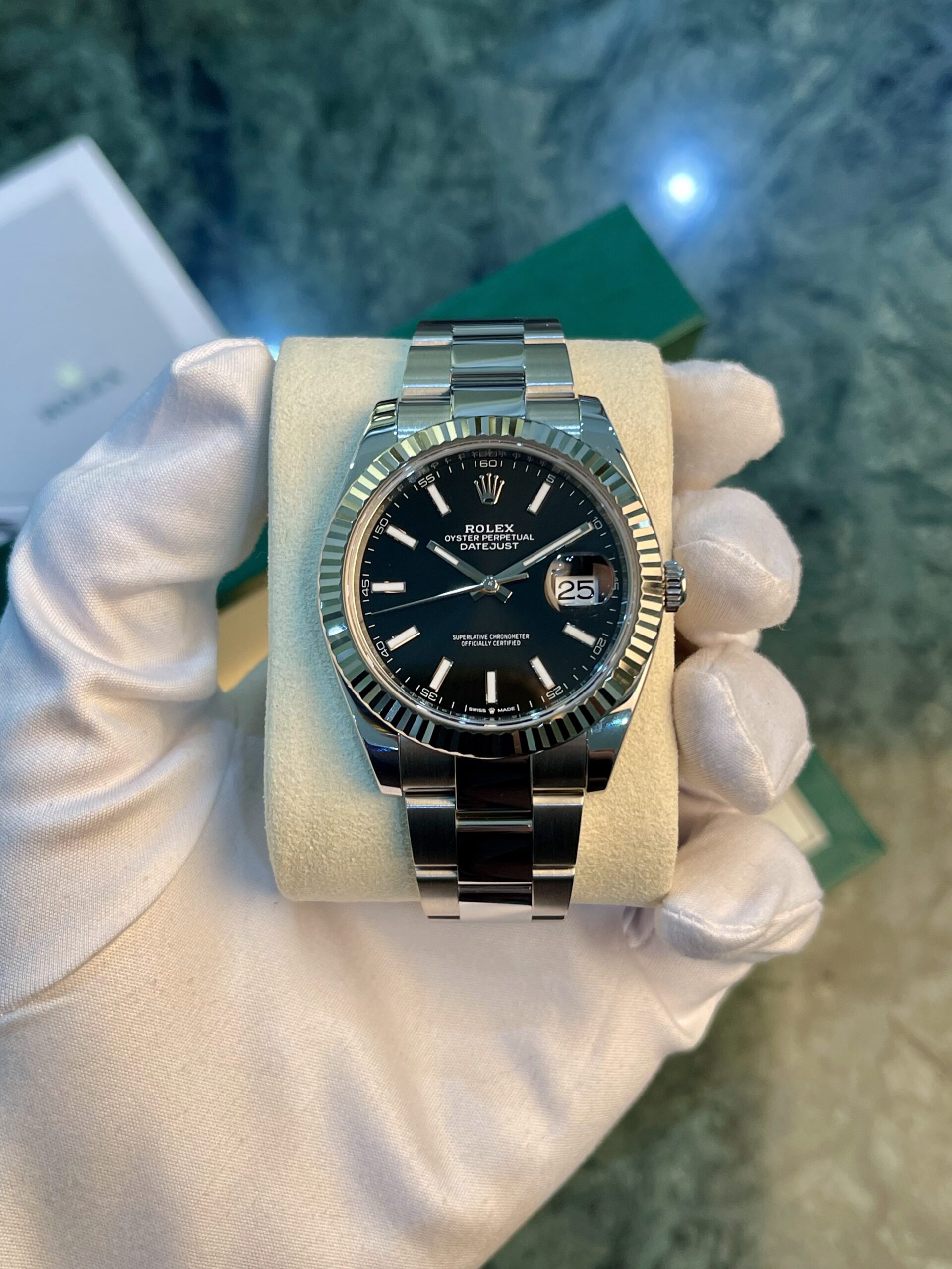 Close-up of a Rolex Oyster Perpetual Datejust stainless steel watch with a black dial and date window, held by a gloved hand