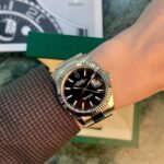 Close-up of a silver Rolex Datejust on a wrist with a black dial and date window, fluted bezel, against a blurred Rolex box in the background.