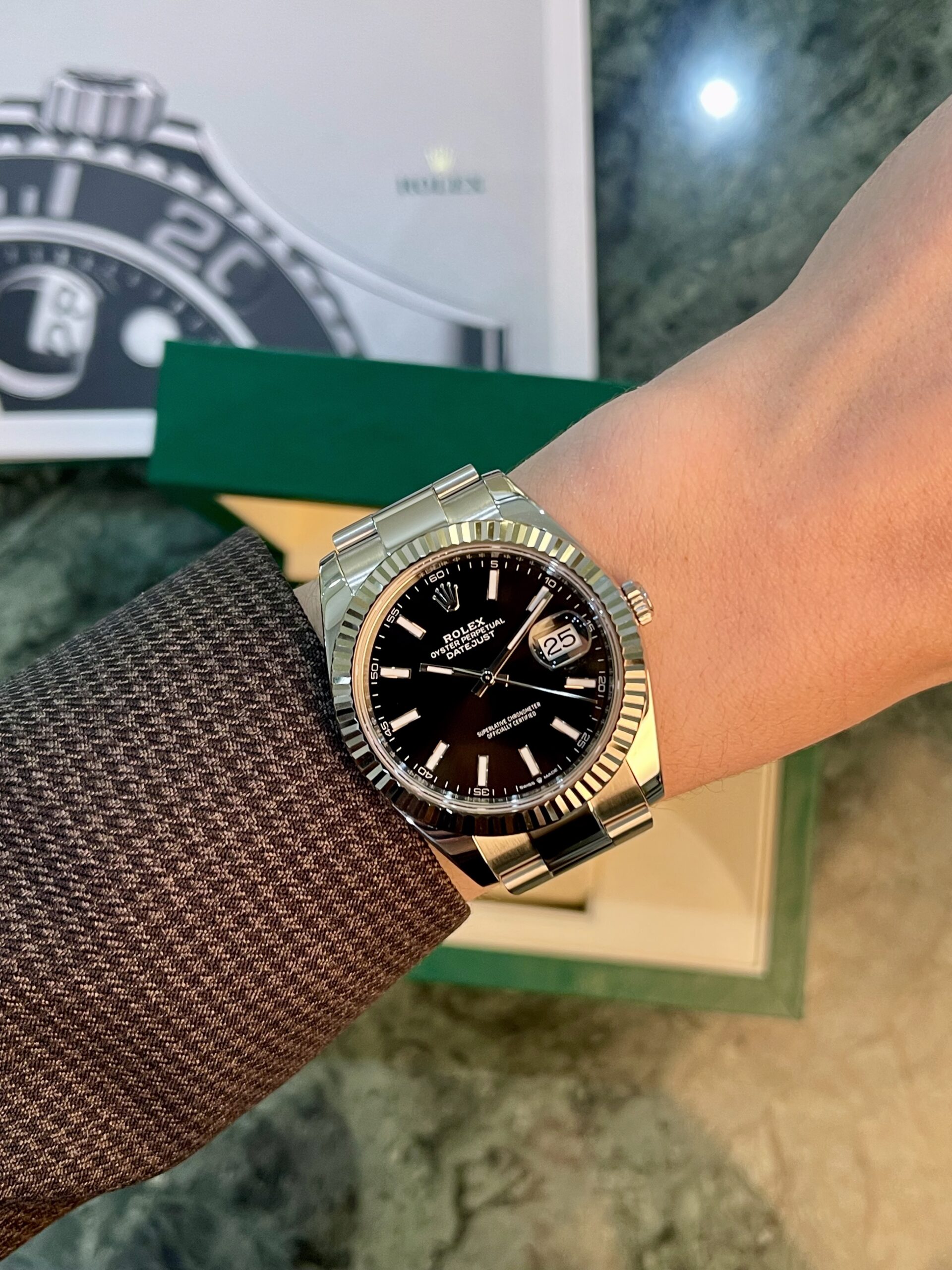 Close-up of a silver Rolex Datejust on a wrist with a black dial and date window, fluted bezel, against a blurred Rolex box in the background.