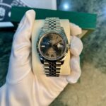 Rolex Datejust wristwatch with Roman numeral dial on a metal bracelet, held by gloved hand on a cushion.