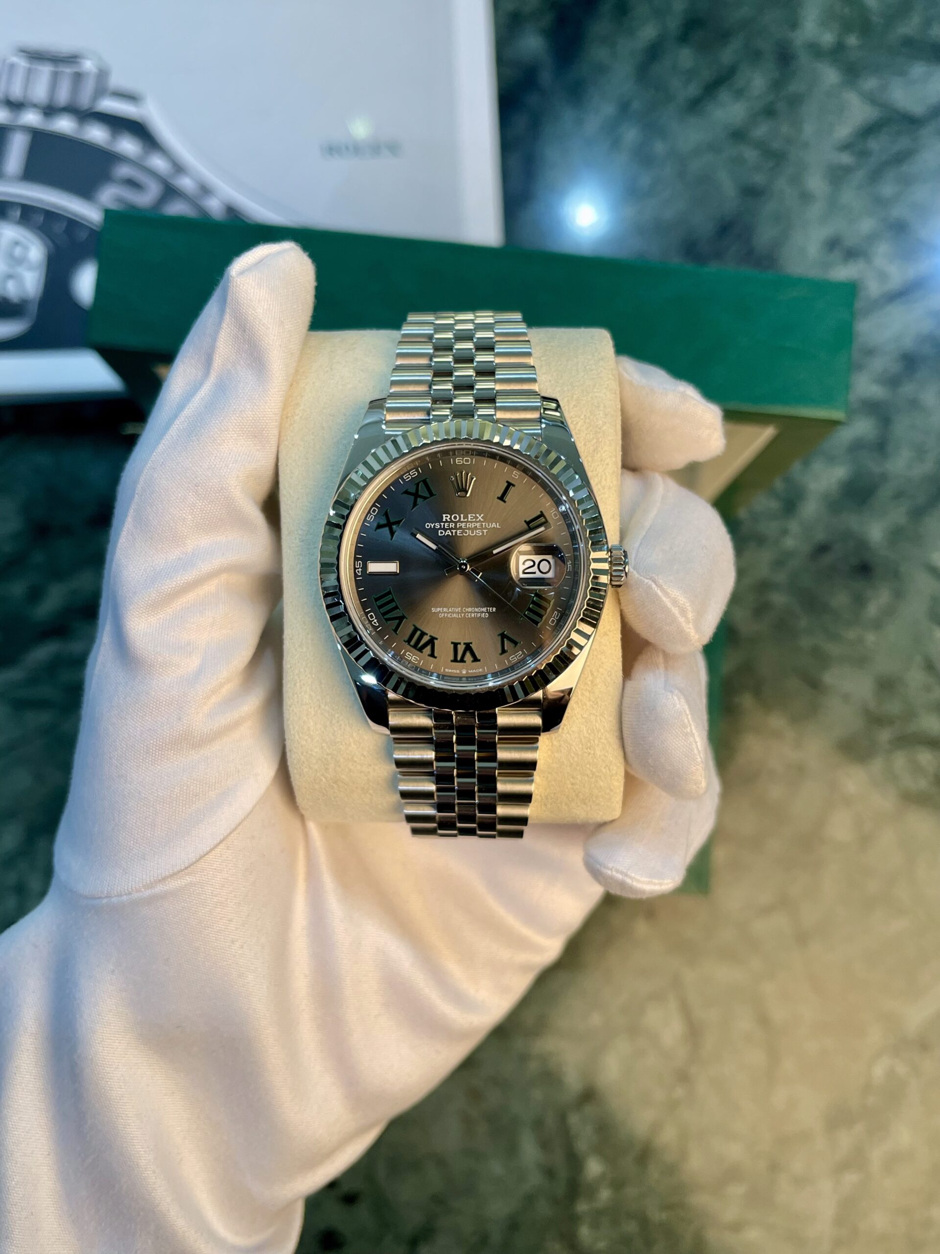 Rolex Datejust wristwatch with Roman numeral dial on a metal bracelet, held by gloved hand on a cushion.