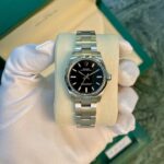 Close-up of a stainless steel Rolex Oyster Perpetual watch on a light display cushion, held by gloved hands in front of a green box edge.