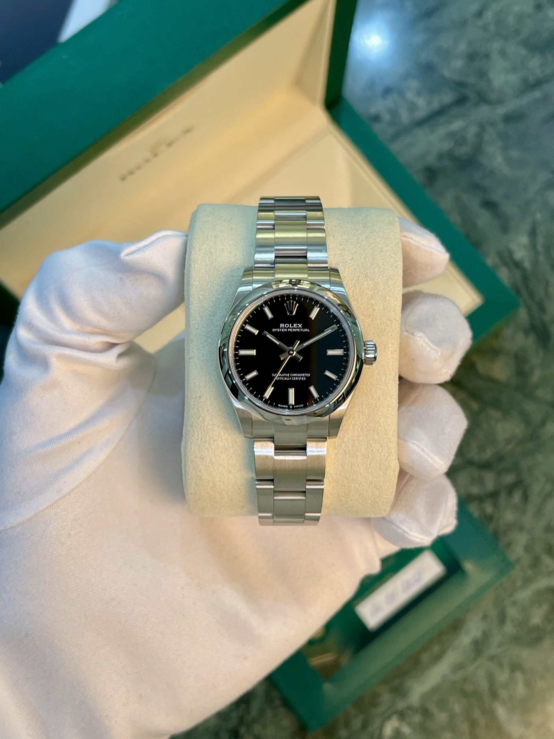 Close-up of a stainless steel Rolex Oyster Perpetual watch on a light display cushion, held by gloved hands in front of a green box edge.