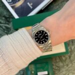 Stainless steel Rolex watch on a beige-sleeved wrist, black dial, with a green Rolex box in the background.