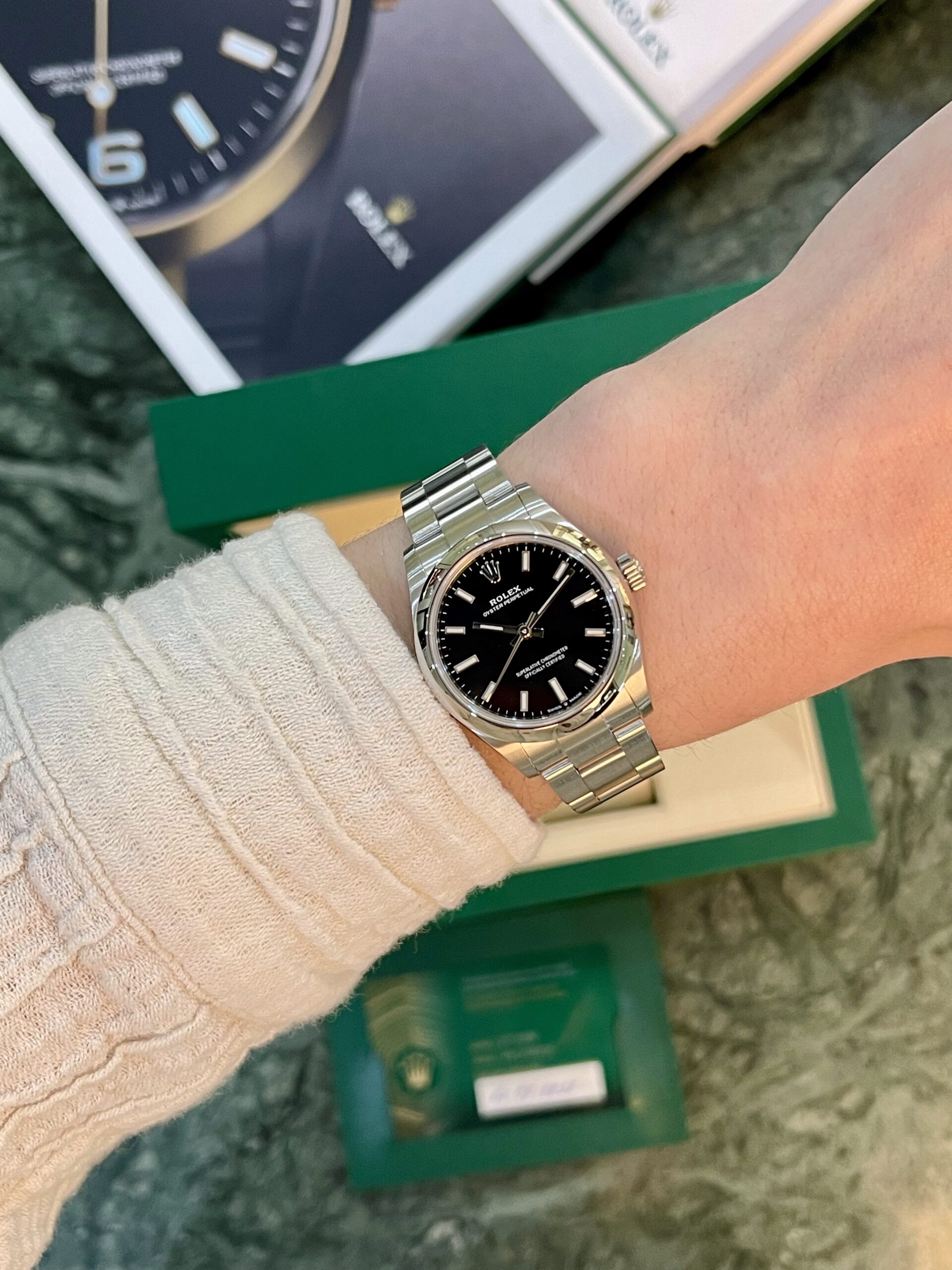 Stainless steel Rolex watch on a beige-sleeved wrist, black dial, with a green Rolex box in the background.