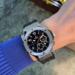 Close-up of a silver stainless steel watch on a blue-sleeved arm, black dial with multiple subdials and white indices, brand visible as Hublot Genève.