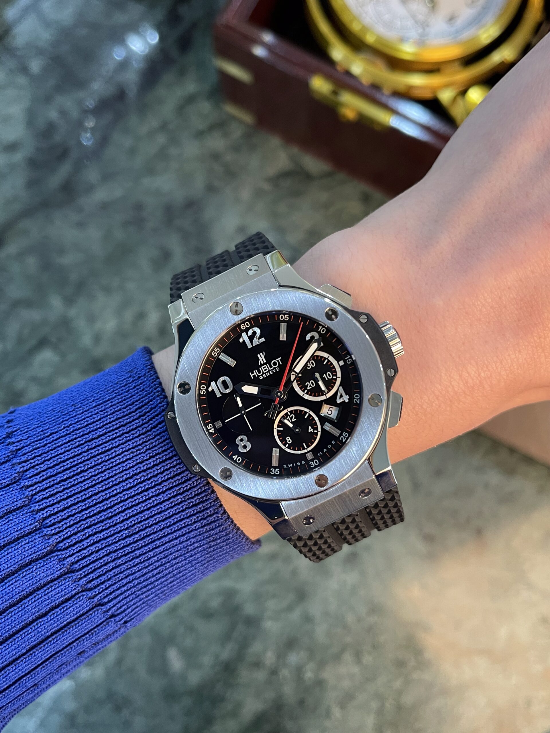 Close-up of a silver stainless steel watch on a blue-sleeved arm, black dial with multiple subdials and white indices, brand visible as Hublot Genève.