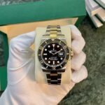 Two-tone Rolex Submariner on a beige cushion, black dial, gold bezel, date window at 3 o'clock, held by gloved hand in front of a green box.