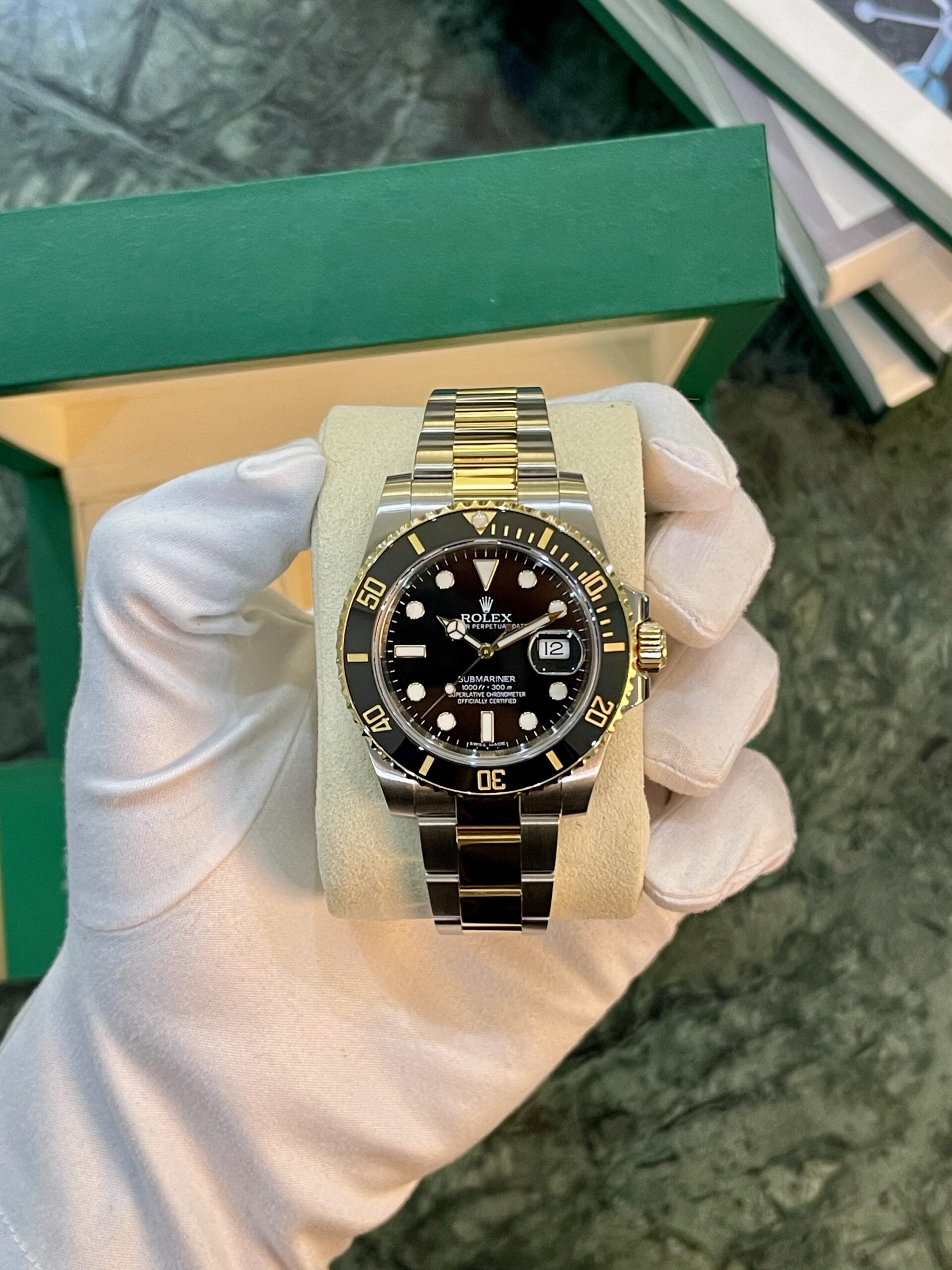 Two-tone Rolex Submariner on a beige cushion, black dial, gold bezel, date window at 3 o'clock, held by gloved hand in front of a green box.