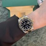 Wrist wearing a two-tone Rolex Submariner with black dial and gold bezel, positioned near a green box.