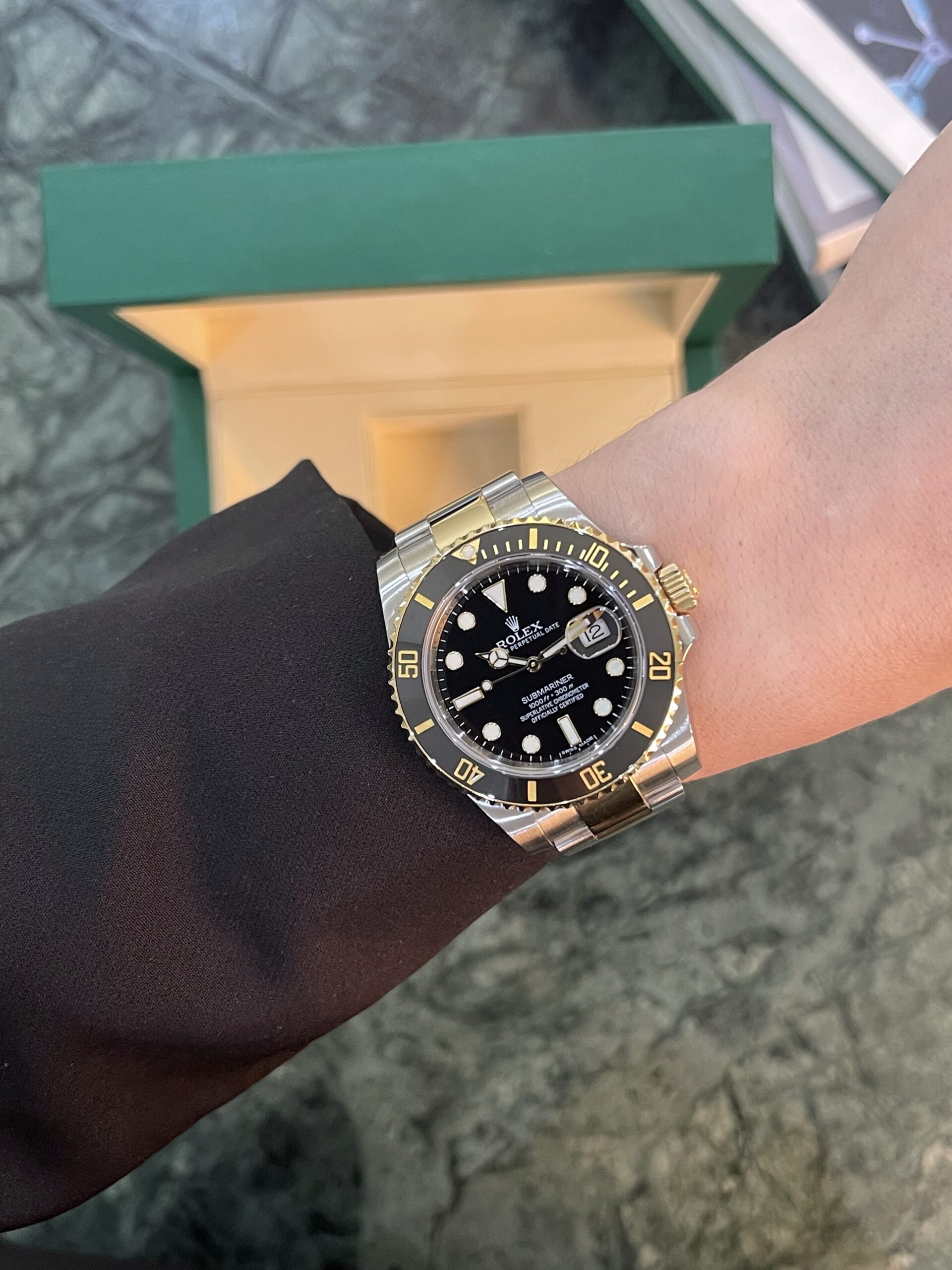 Wrist wearing a two-tone Rolex Submariner with black dial and gold bezel, positioned near a green box.