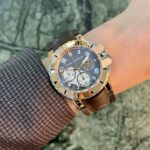 Gold-toned luxury watch with brown strap on the wrist, partial gray textured sleeve, green outdoor background.