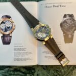 Gold-tone wristwatch with a black strap resting on an open catalog page showing Ocean Dual Time watches against a green marble surface.