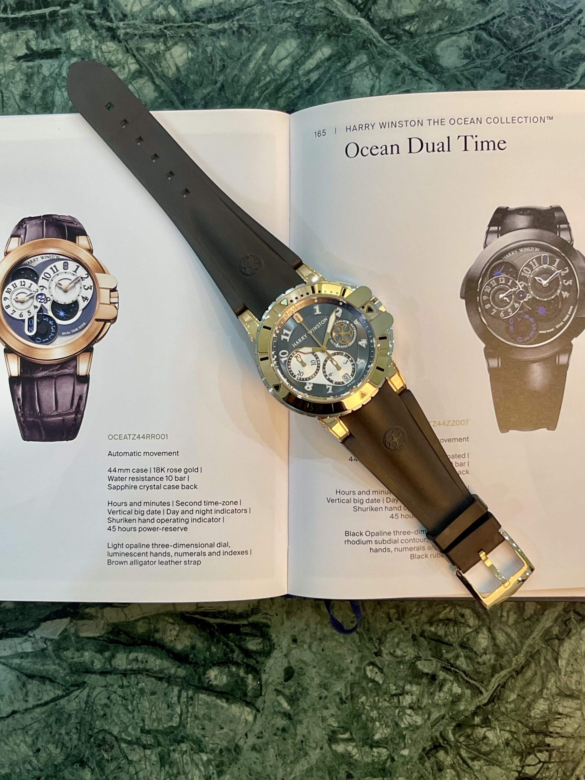 Gold-tone wristwatch with a black strap resting on an open catalog page showing Ocean Dual Time watches against a green marble surface.