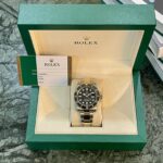 Two-tone Rolex dive watch in a green presentation box with cream interior and warranty card nearby on a marble surface
