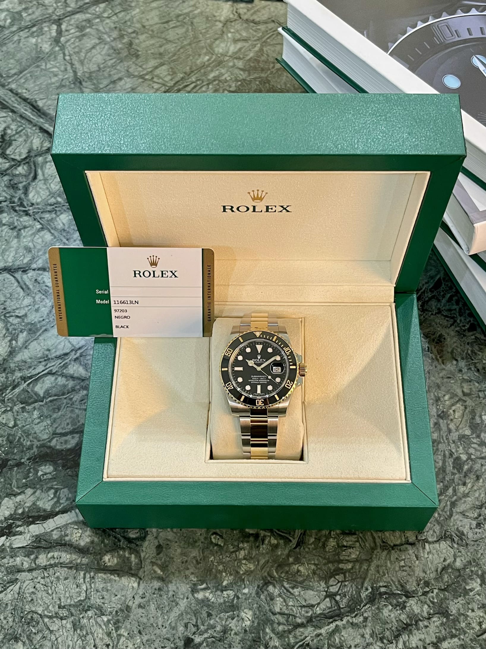 Two-tone Rolex dive watch in a green presentation box with cream interior and warranty card nearby on a marble surface