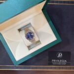 Green jewelry box opened to reveal a two-tone metal wristwatch with a blue dial inside a cream interior. A Privilegia card is nearby.