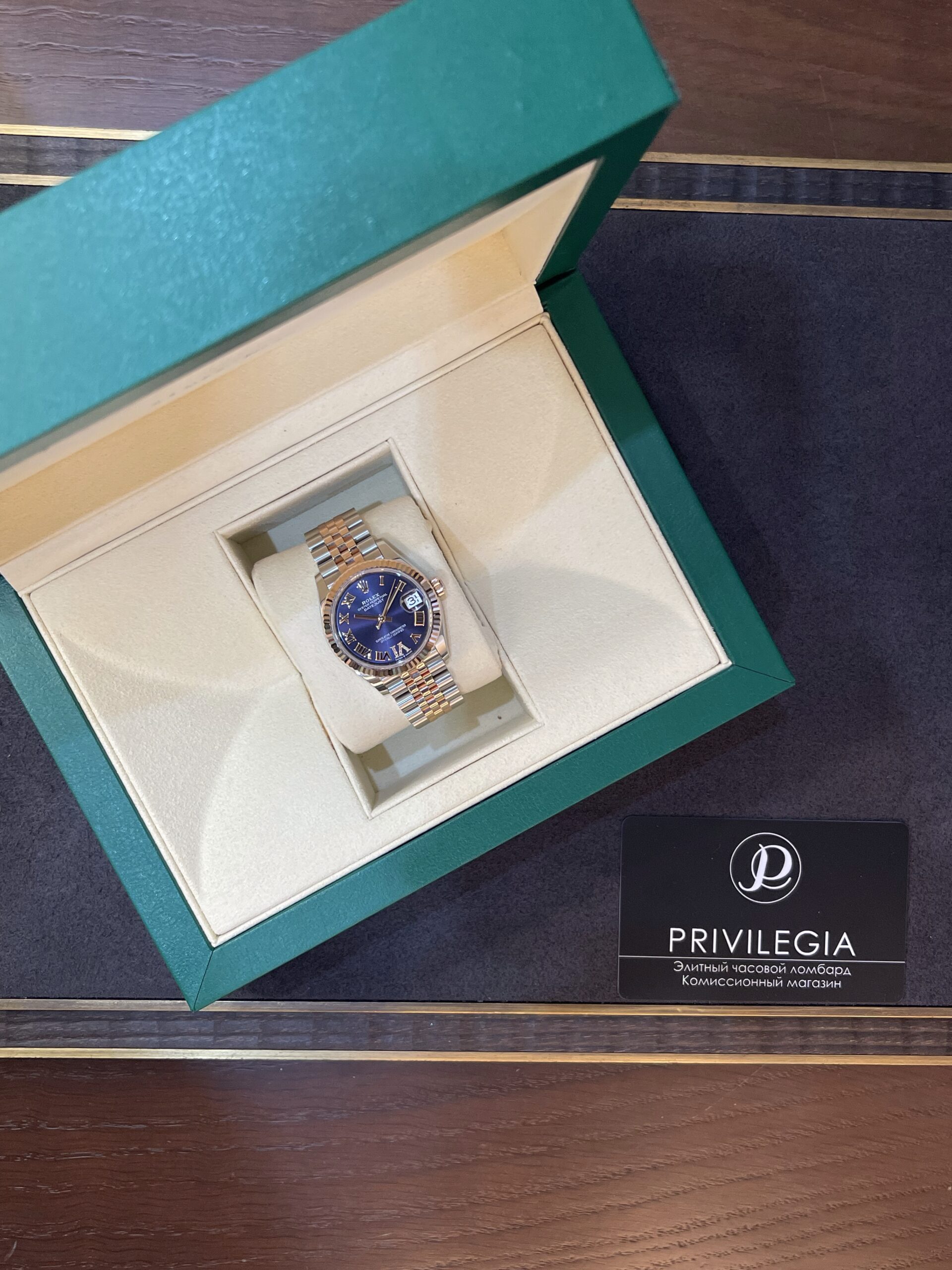 Green jewelry box opened to reveal a two-tone metal wristwatch with a blue dial inside a cream interior. A Privilegia card is nearby.