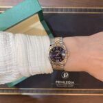 Wrist wearing a two-tone Rolex Datejust with a blue dial, next to a green textured box.