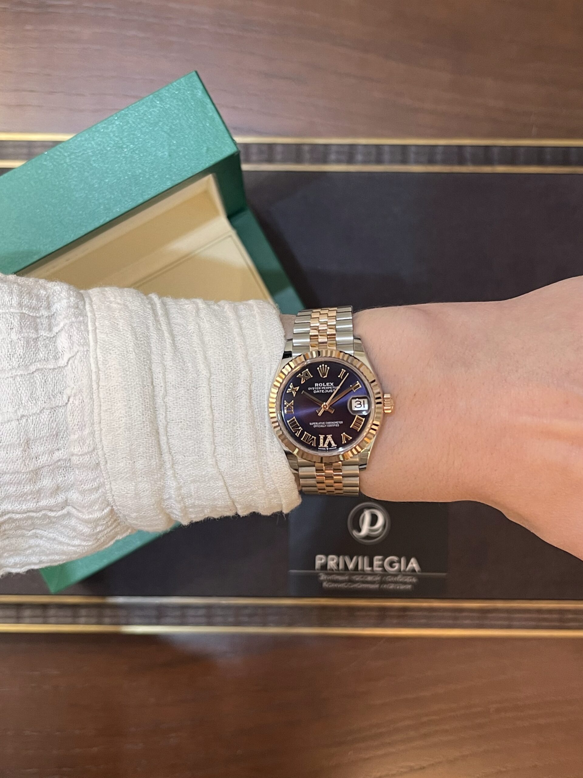 Wrist wearing a two-tone Rolex Datejust with a blue dial, next to a green textured box.