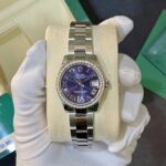 Rolex pearl-white-diamonds bezel watch with a purple dial in a cream display cushion, held by gloved hands against a green box background. (informative)