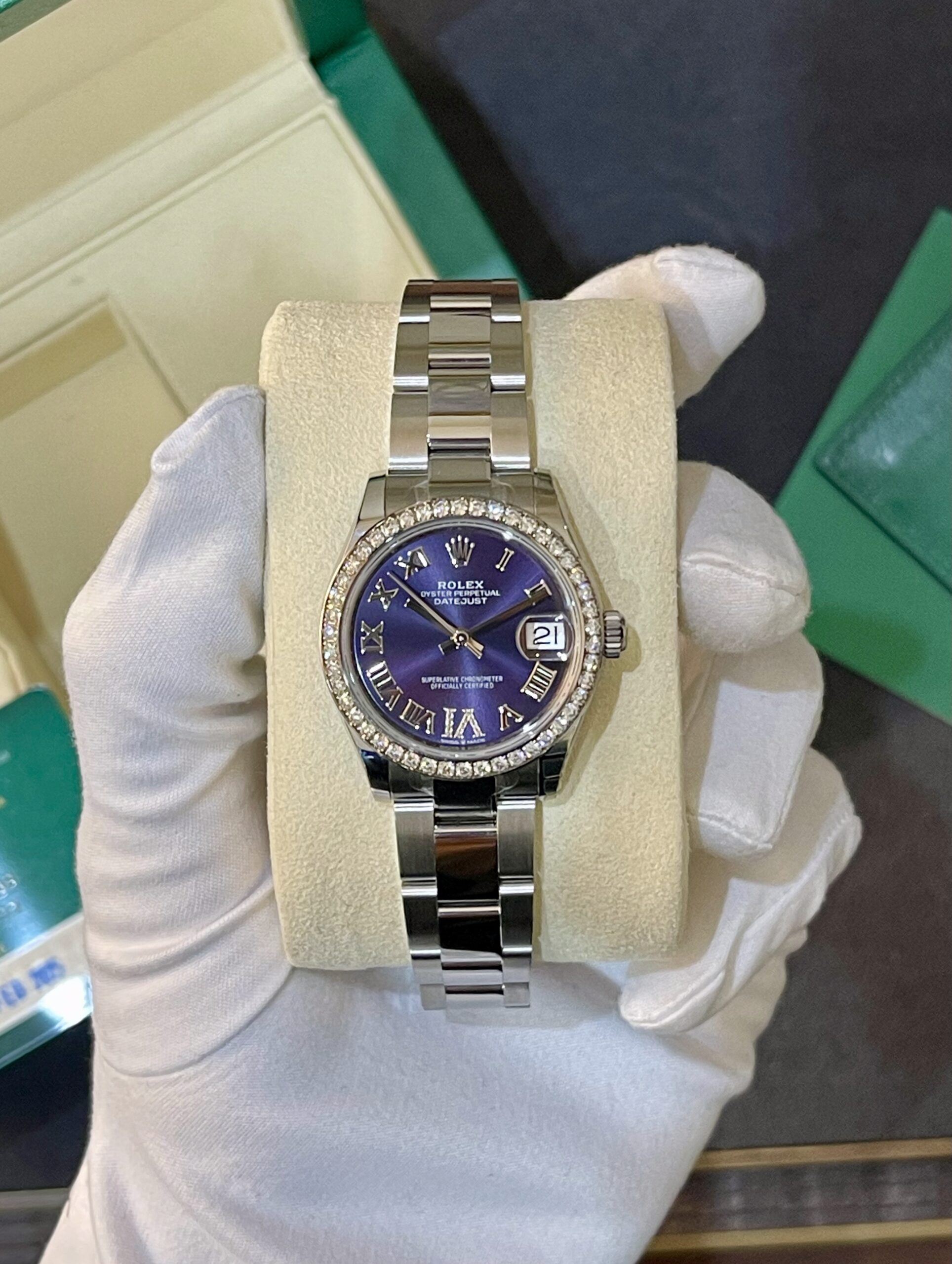 Rolex pearl-white-diamonds bezel watch with a purple dial in a cream display cushion, held by gloved hands against a green box background. (informative)
