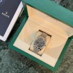 Rolex stainless steel watch displayed in a beige cushion inside a green presentation box on a dark marble surface, ready for viewing.