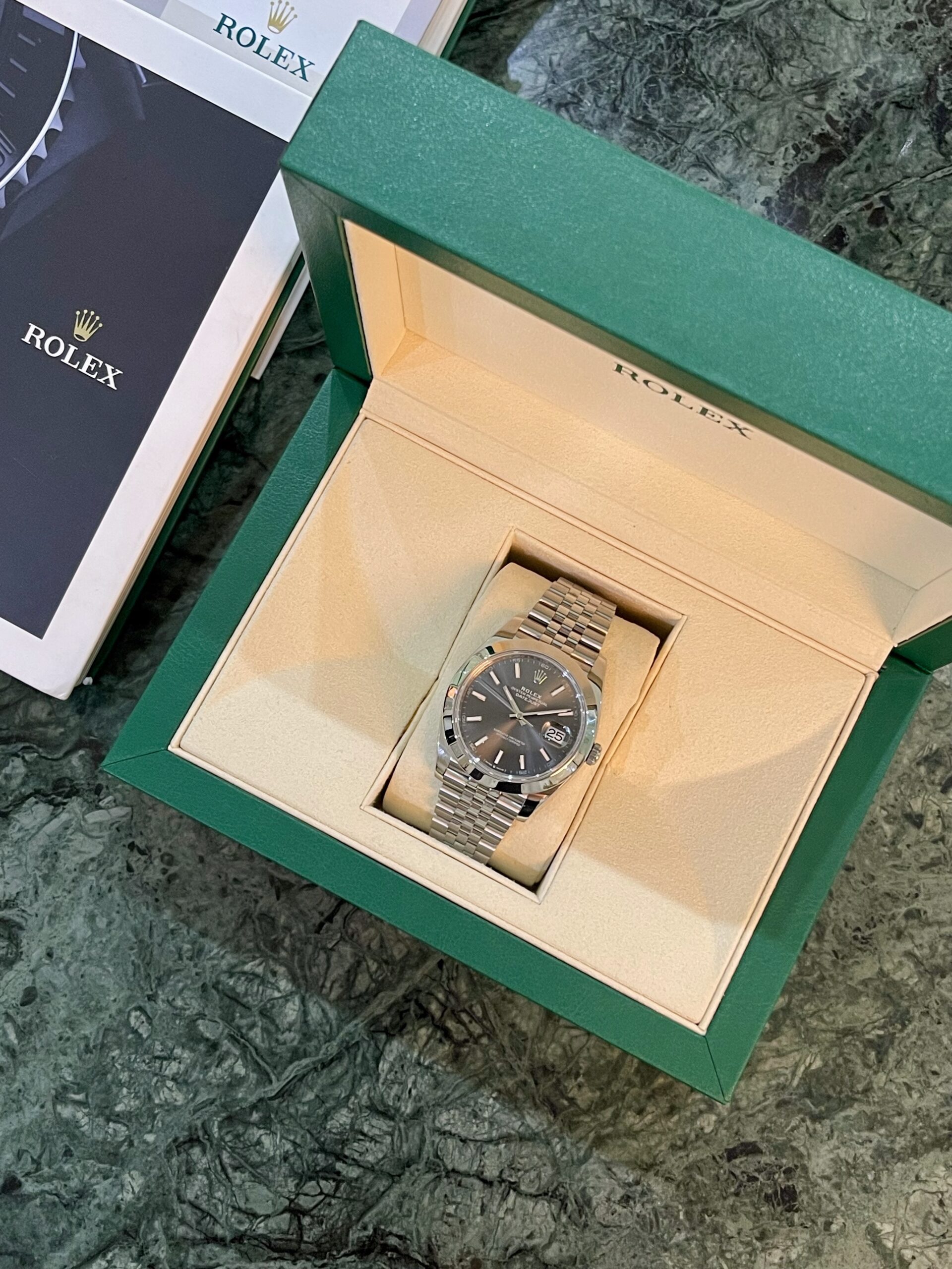 Rolex stainless steel watch displayed in a beige cushion inside a green presentation box on a dark marble surface, ready for viewing.