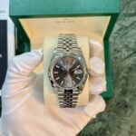 Silver Rolex Datejust wristwatch on a beige cushion, held by a white-gloved hand with a green box in the background, front view.