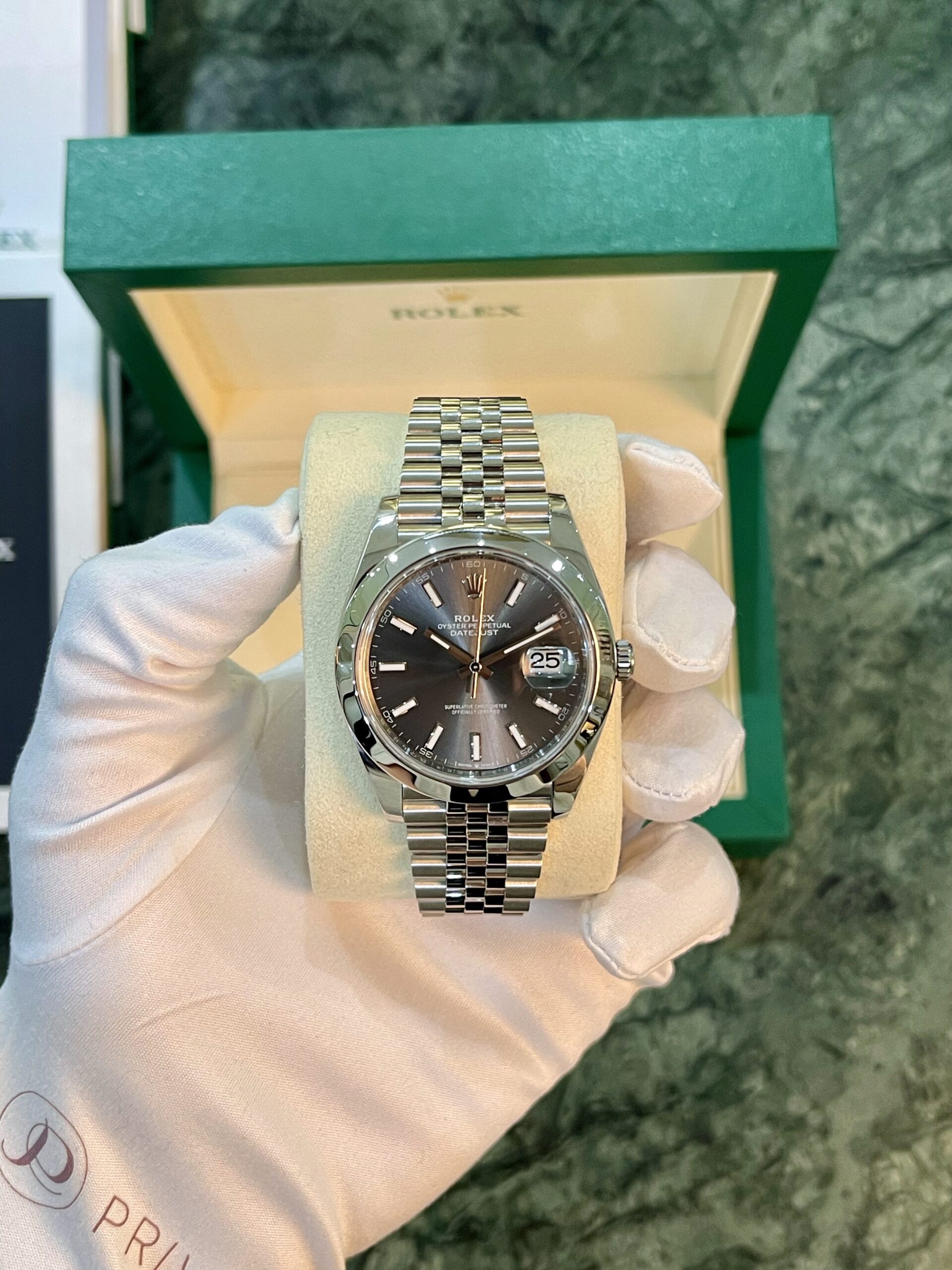 Silver Rolex Datejust wristwatch on a beige cushion, held by a white-gloved hand with a green box in the background, front view.