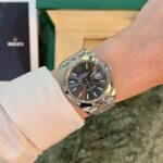 Wrist with a silver Rolex Datejust watch and a dark dial, showing the date at 3 o'clock.