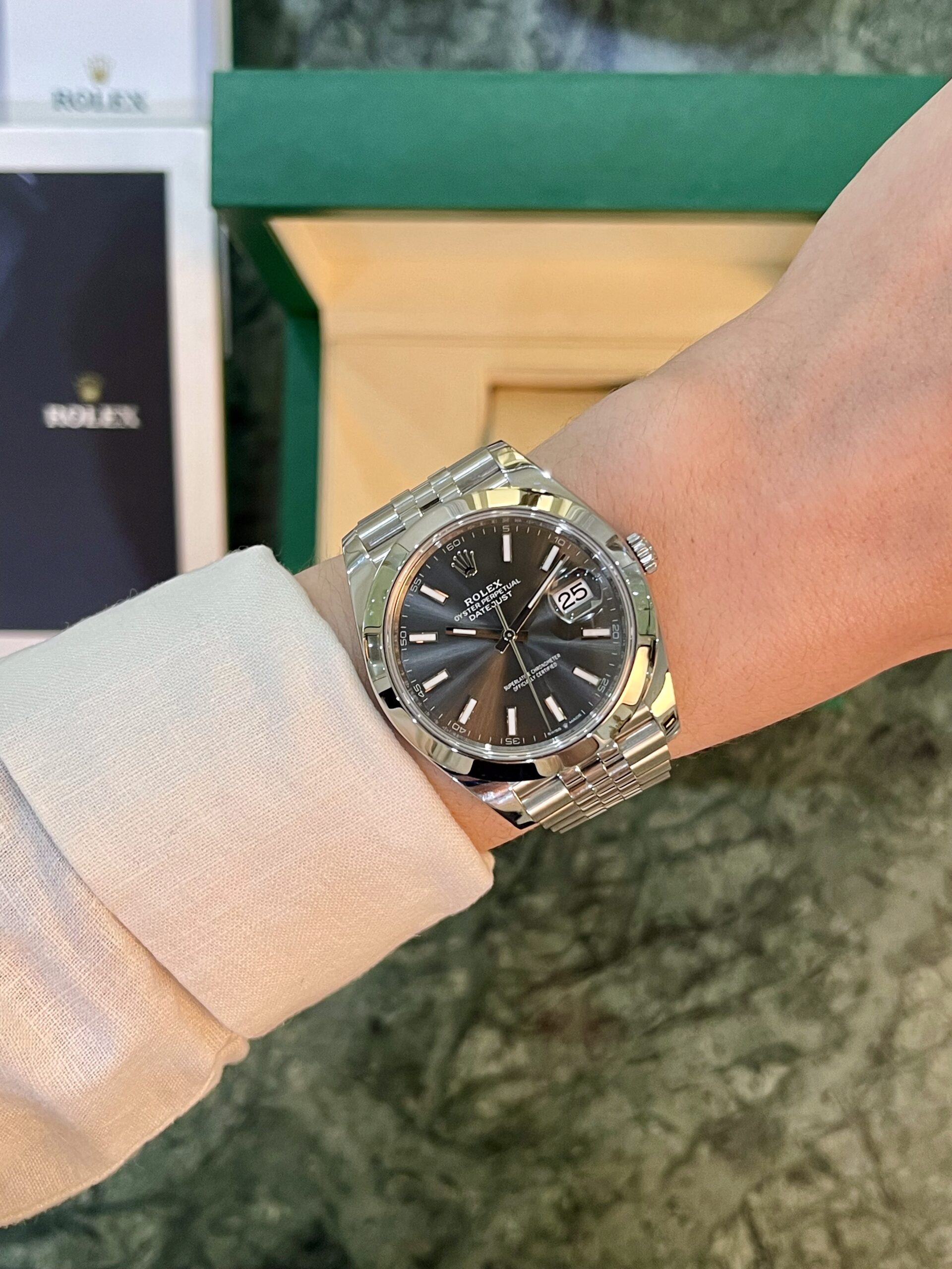 Wrist with a silver Rolex Datejust watch and a dark dial, showing the date at 3 o'clock.