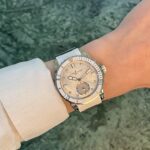 Wristwatch on a forearm: two-tone metal watch with a beige dial, diamond markers, and Ulysse Nardin branding, worn over a light-colored sleeve with a green marbled background