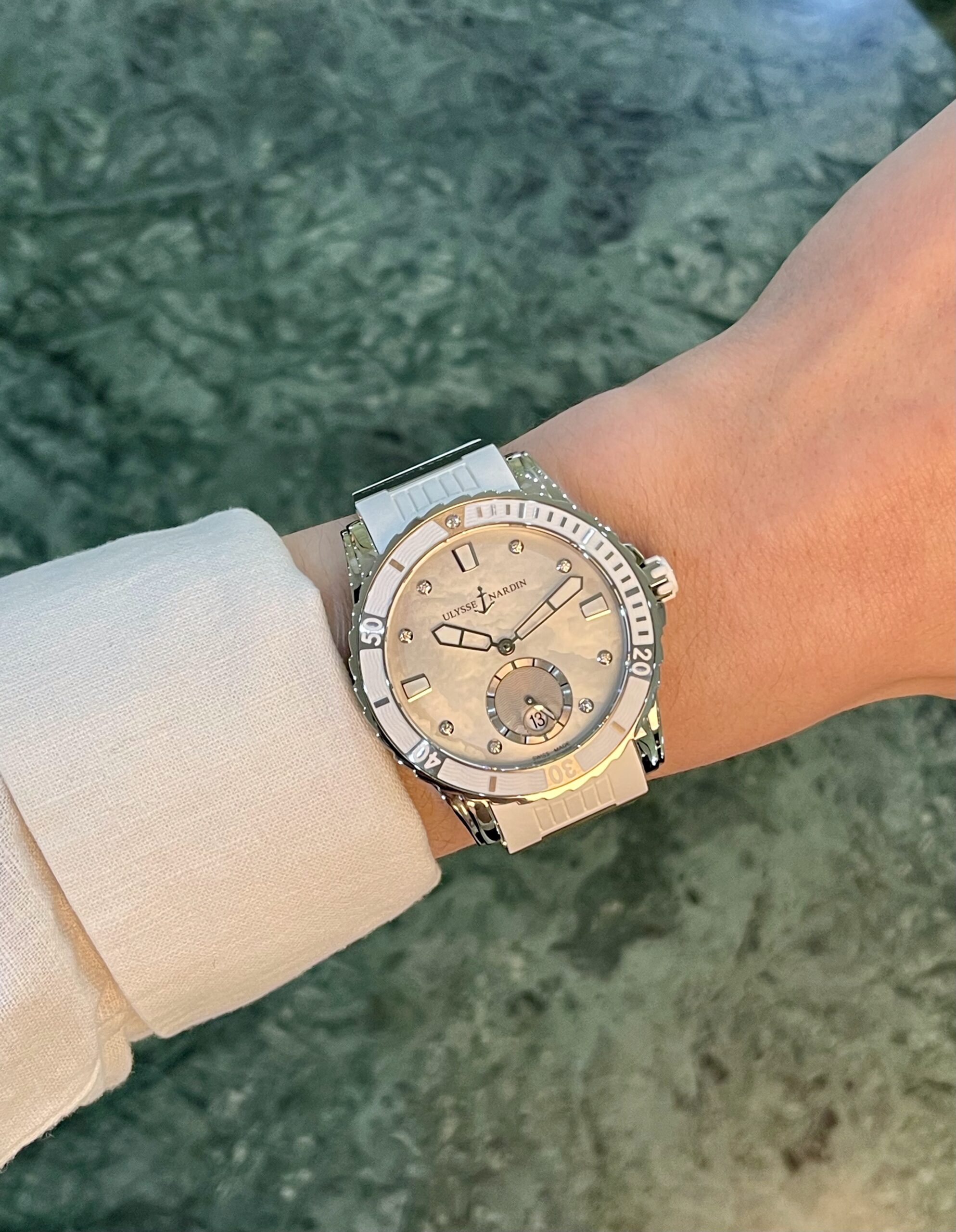 Wristwatch on a forearm: two-tone metal watch with a beige dial, diamond markers, and Ulysse Nardin branding, worn over a light-colored sleeve with a green marbled background