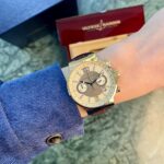 Gold Ulysse Nardin wristwatch worn on a blue-sleeved wrist with a red Ulysse Nardin box in the background on a marble-like floor.
