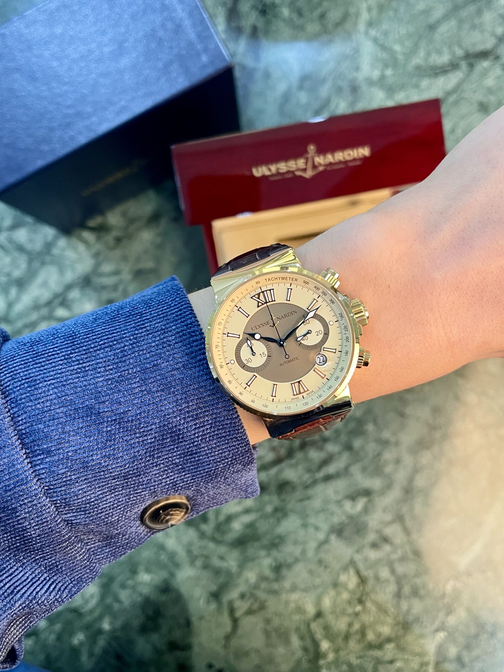 Gold Ulysse Nardin wristwatch worn on a blue-sleeved wrist with a red Ulysse Nardin box in the background on a marble-like floor.