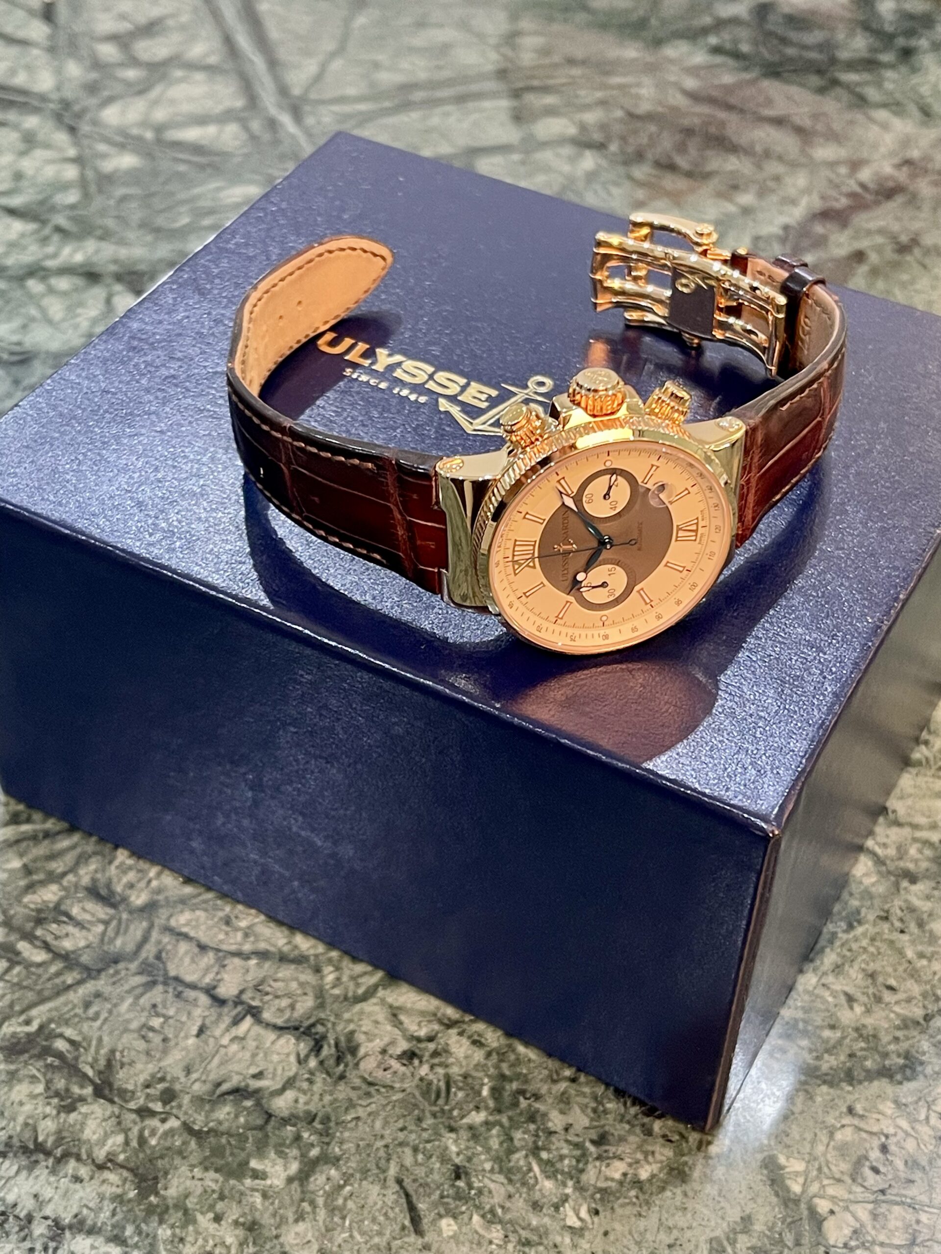 Gold-tone chronograph wristwatch with a brown leather strap resting on a dark blue ULYSSE box, partially showing the brand name and anchor logo.