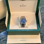 Rolex watch in an open green presentation box, silver bracelet and blue dial visible inside beige padding