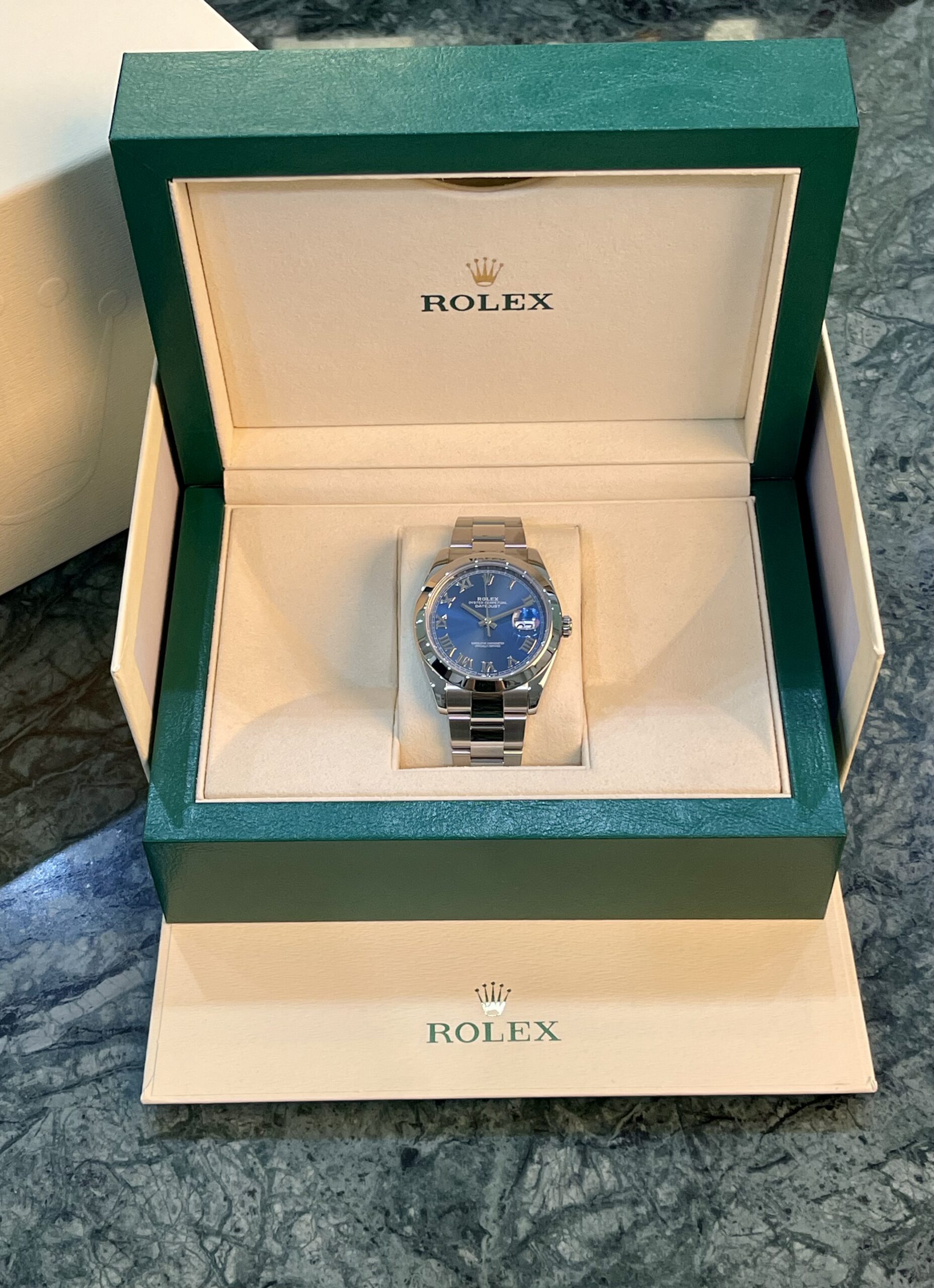 Rolex watch in an open green presentation box, silver bracelet and blue dial visible inside beige padding
