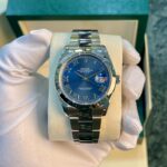 Rolex Oyster Perpetual Datejust watch with blue dial and Roman numerals held by gloved hand in a box cushion.