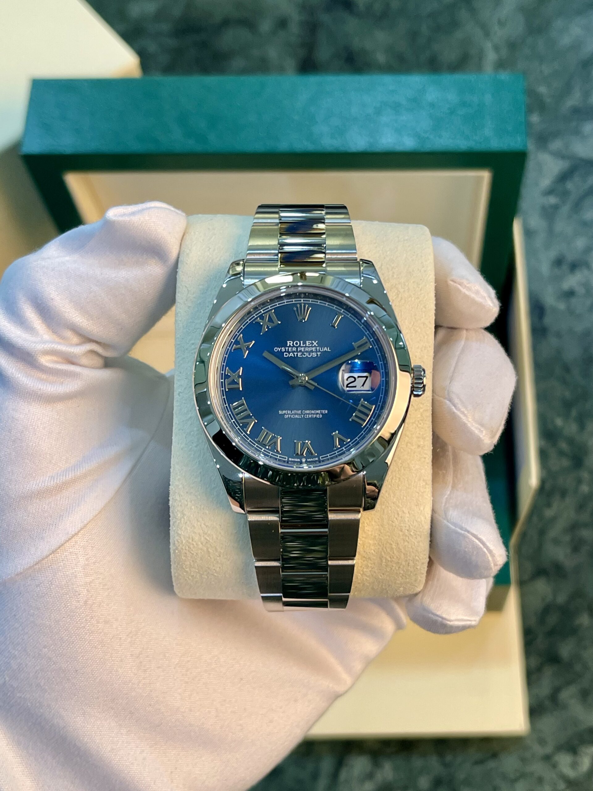 Rolex Oyster Perpetual Datejust watch with blue dial and Roman numerals held by gloved hand in a box cushion.