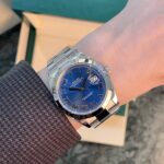 Wristwatch with a blue dial and silver metal band on a forearm, with a green box in the background.