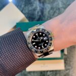 Rolex GMT-Master II on the wrist with a black dial, gold-and-black bezel, and stainless steel bracelet over a textured sleeve.