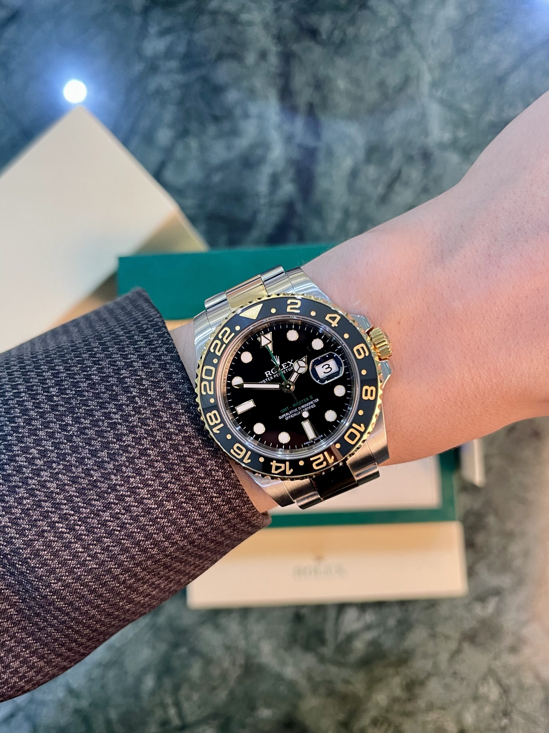 Rolex GMT-Master II on the wrist with a black dial, gold-and-black bezel, and stainless steel bracelet over a textured sleeve.