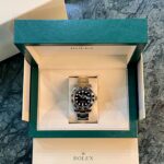 Open Rolex presentation box with green outer lid, beige interior, housing a silver Rolex watch with black dial and rotating bezel on a marble surface; Rolex logo visible inside and on bottom box.