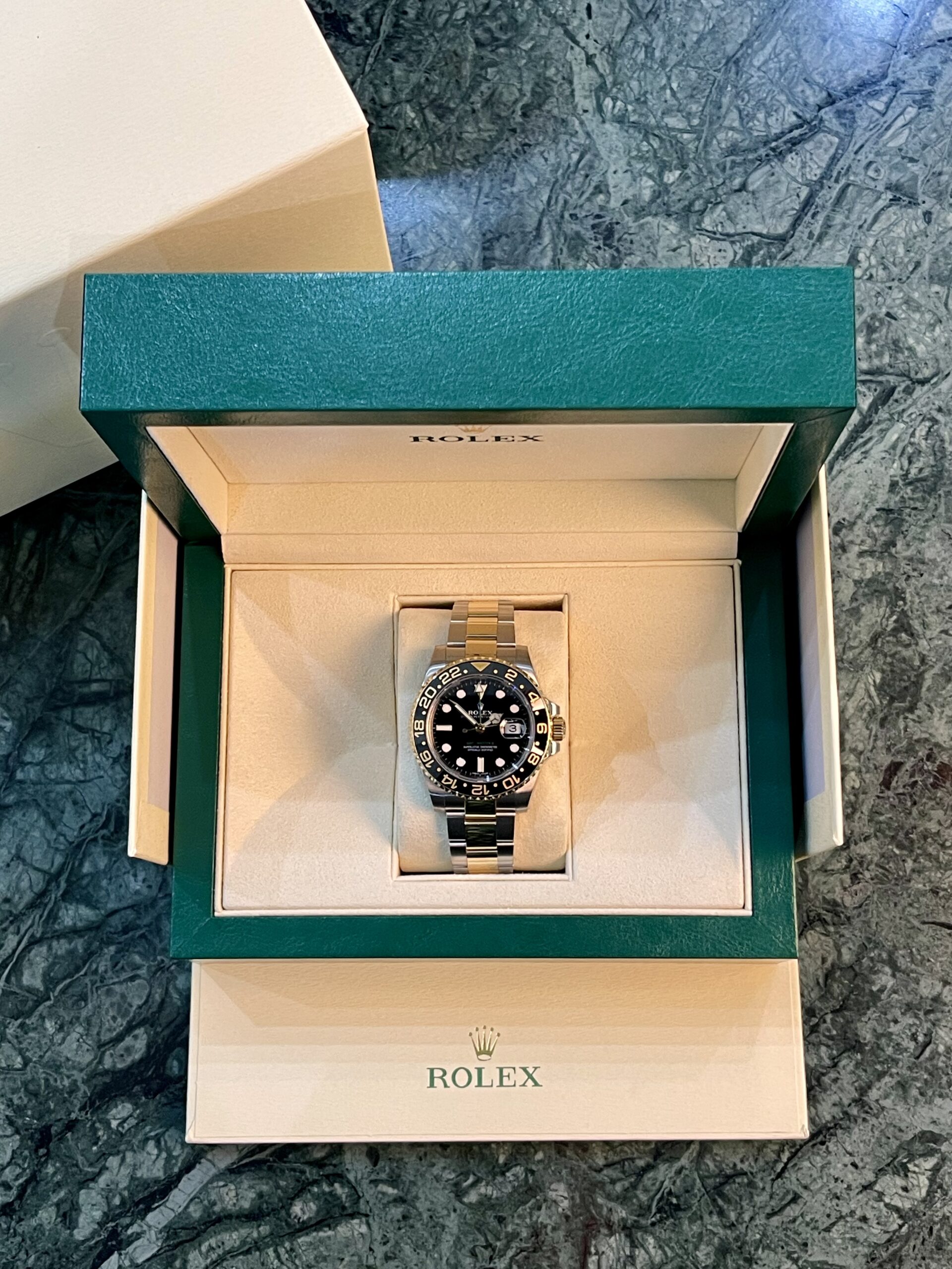 Open Rolex presentation box with green outer lid, beige interior, housing a silver Rolex watch with black dial and rotating bezel on a marble surface; Rolex logo visible inside and on bottom box.