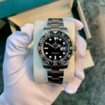 Rolex GMT‑Master II watch held in a gloved hand, stainless steel bracelet, black dial, gold accents, in a box.