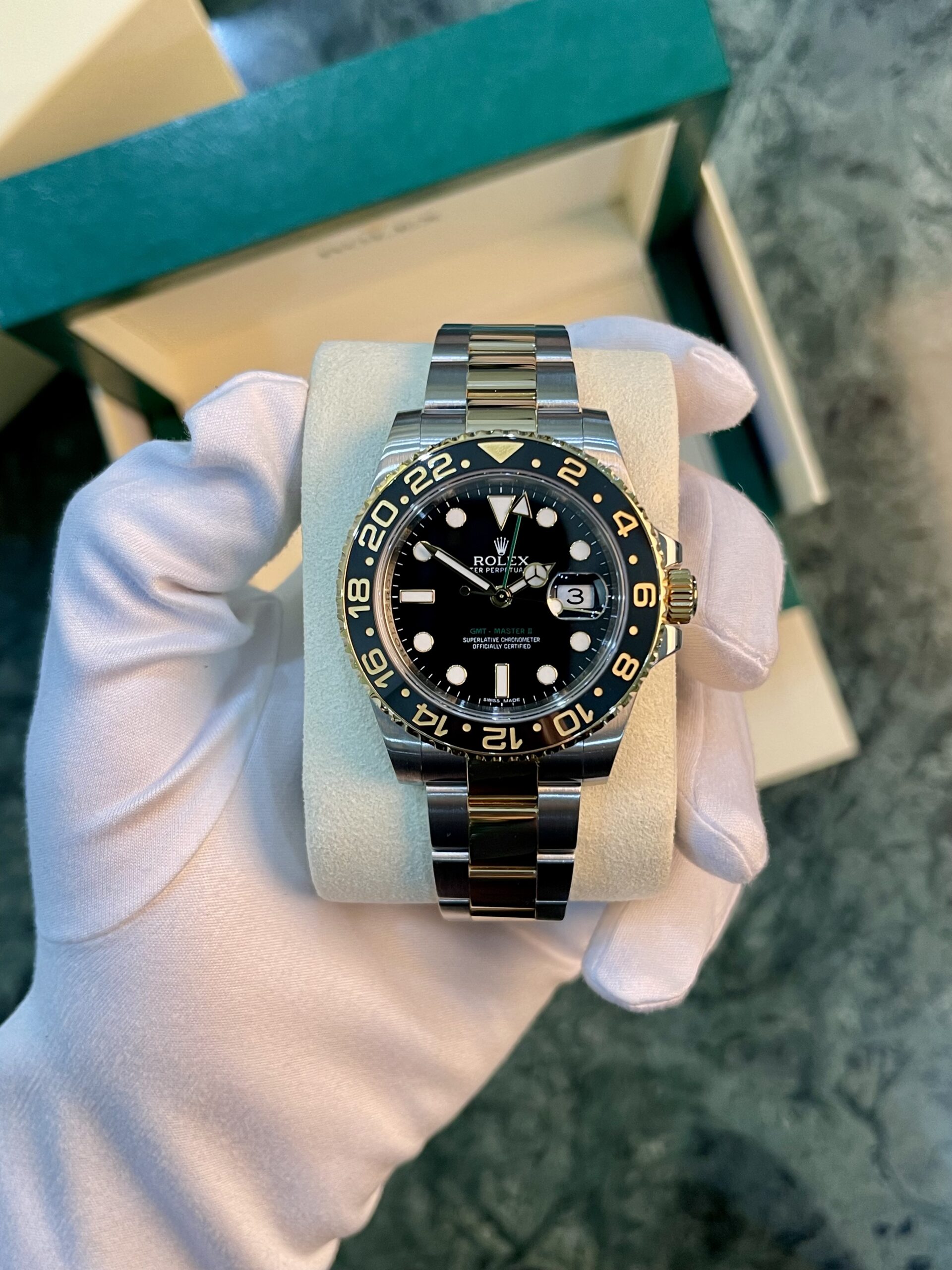 Rolex GMT‑Master II watch held in a gloved hand, stainless steel bracelet, black dial, gold accents, in a box.