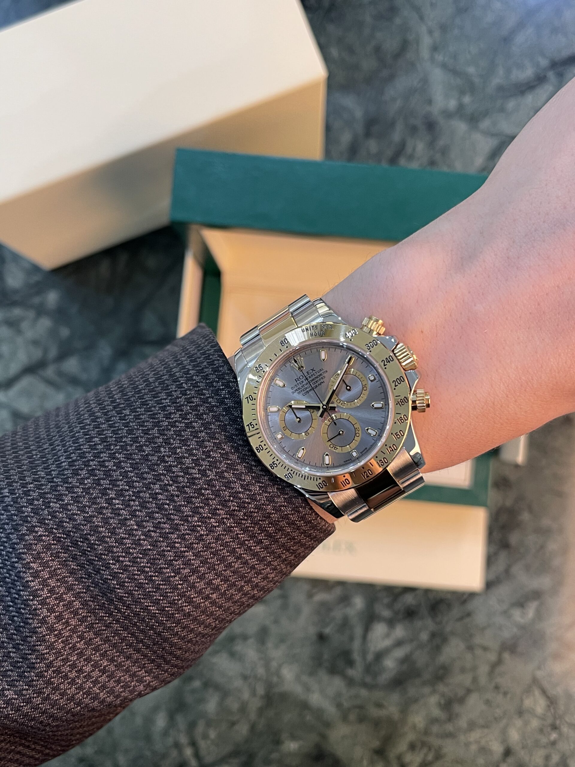 Two-tone Rolex watch on a wrist with a denim sleeve and beige packaging boxes in the background.