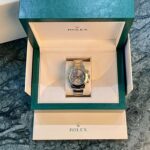 Open Rolex presentation box with a two-tone Rolex watch on a cushioned insert; green outer box and logo visible on marble surface